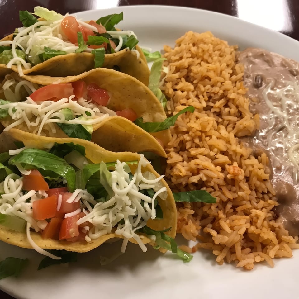 Plate of three hard-shell tacos with lettuce, tomatoes, and shredded cheese, served with Mexican rice and refried beans.