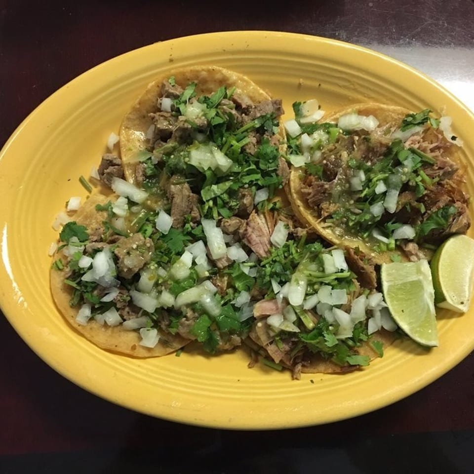 Three Mexican tacos topped with chopped onion, cilantro, green salsa, and meat, served with lime wedges on a yellow plate.