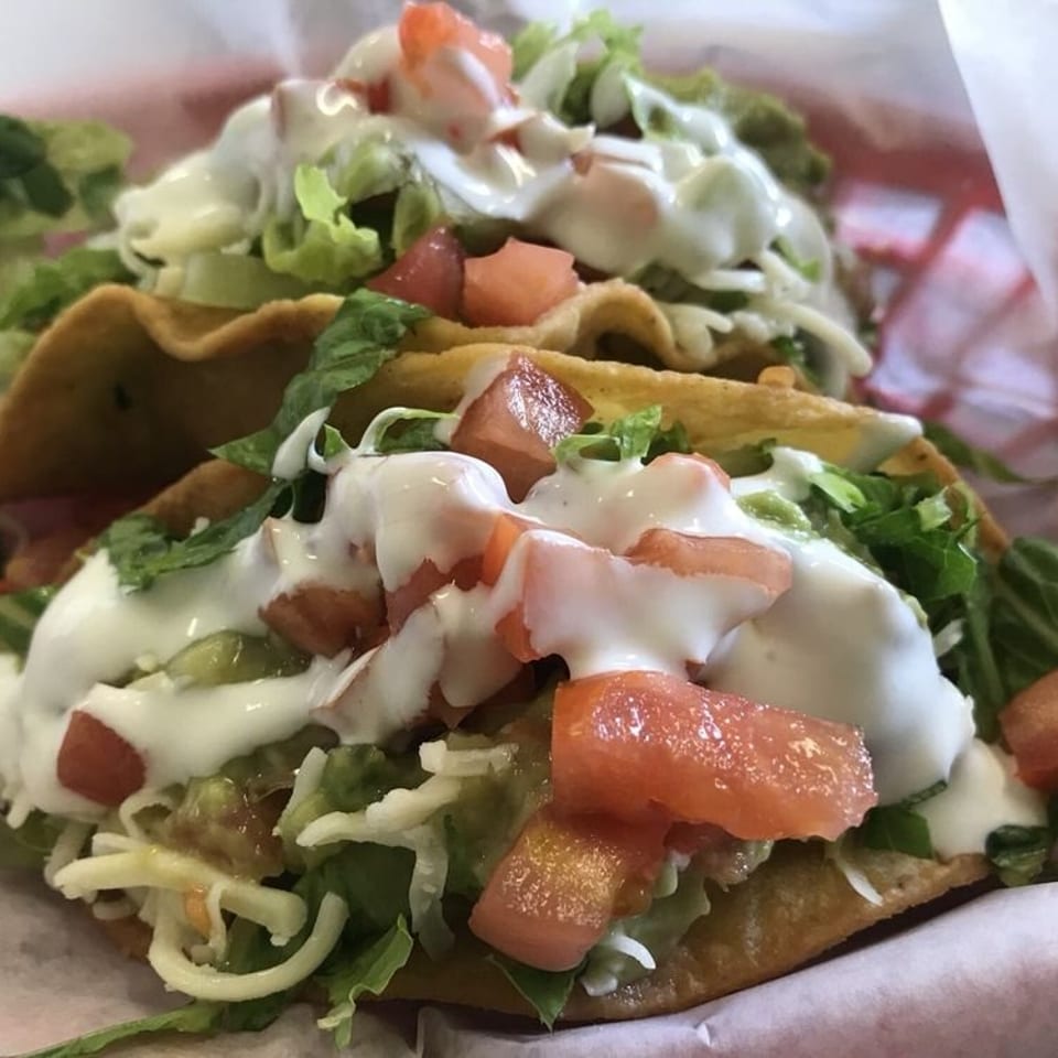 Two crispy tacos topped with lettuce, shredded cheese, diced tomatoes, guacamole, and creamy white sauce.